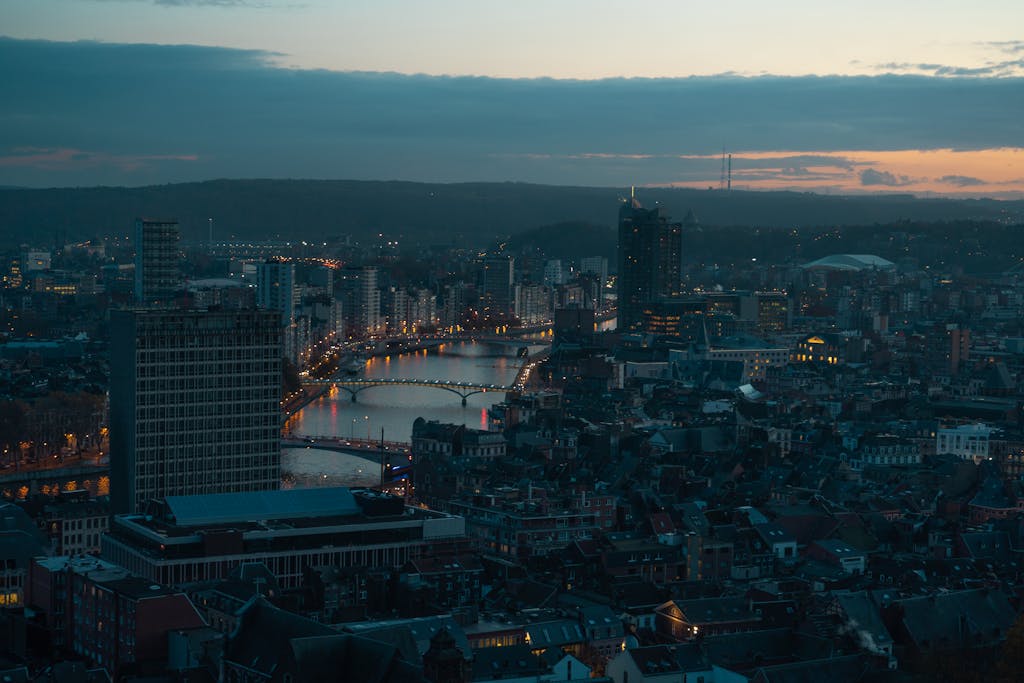 Bridges above broad river surrounded by small houses and tall constructions in megapolis under light pink sky covered with clouds in evening